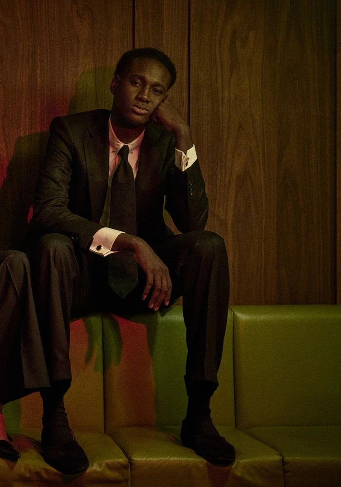 Model Aliou Gueye in a dark suit and pink collared shirt with a tie, seated on a green bench against a wood-paneled wall.