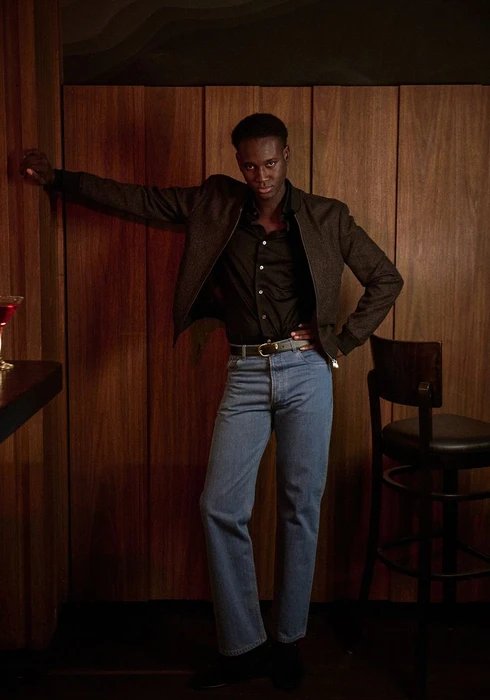 Model leaning against a wood panel wall wearing a dark jacket, black shirt, and blue jeans in a bar setting.