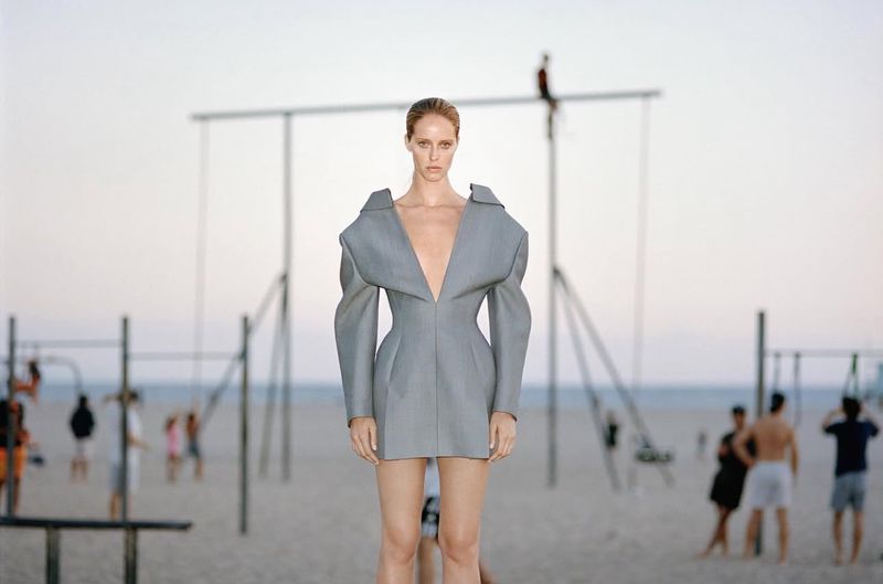 Fashion shot of a slim adult woman in a grey dress on a beach with other people and a swing set in the background on a sunny day.