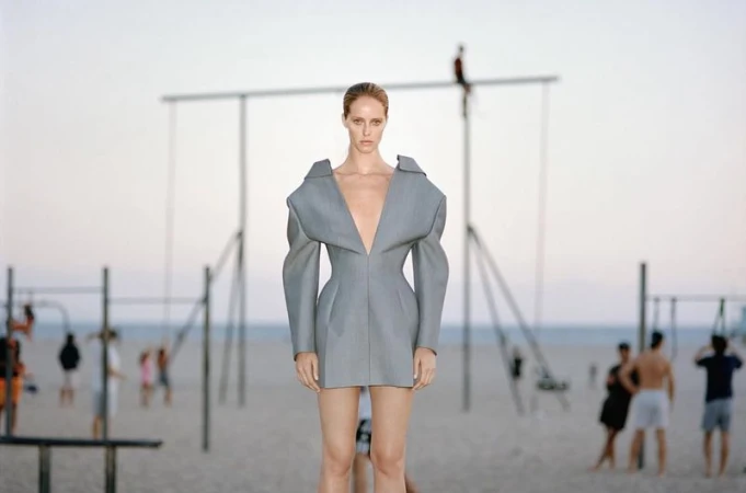 Fashion shot of a slim adult woman in a grey dress on a beach with other people and a swing set in the background on a sunny day.