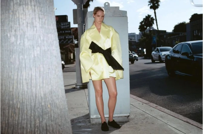 Fashion shot of a tall woman in a yellow coat with a black belt, standing on a city sidewalk with cars and buildings in the background.