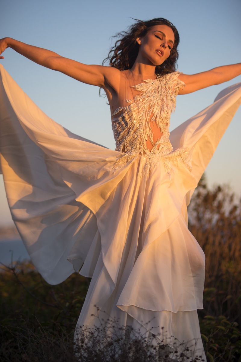 Woman in flowing white gown with feathered detail posing outdoors at sunset with arms outstretched in a dance-like motion.