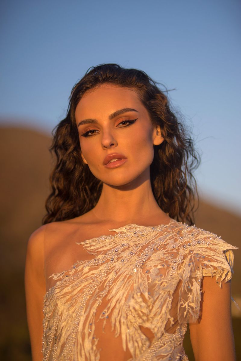 Brunette woman in one-shoulder gown with feathered embellishments poses against a blurred mountain and blue sky backdrop.