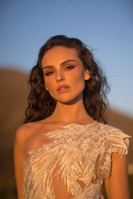 Brunette woman in one-shoulder gown with feathered embellishments poses against a blurred mountain and blue sky backdrop.