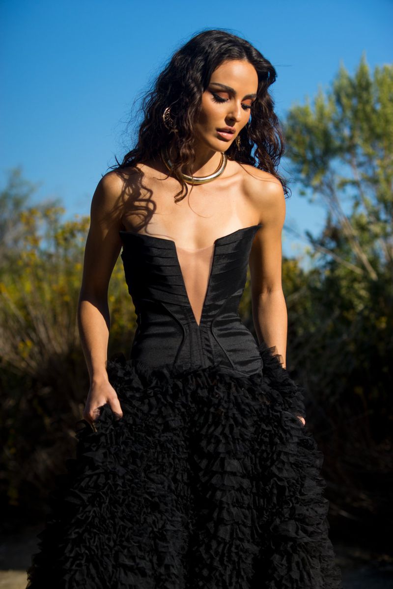 A woman in a black strapless dress poses outdoors in a field with greenery and a clear blue sky, looking down with a serene expression.