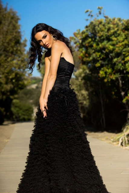 Elegant woman in black strapless gown posing outdoors on path surrounded by lush greenery under a bright blue sky in natural light.