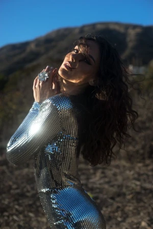 Woman in a sparkling silver sequin jumpsuit with a large cocktail ring posing outdoors against a mountain background.