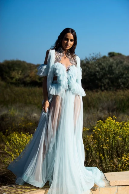 Brunette woman in a light blue ruffled gown stands outdoors against a clear blue sky and natural grassy background, fashion style.