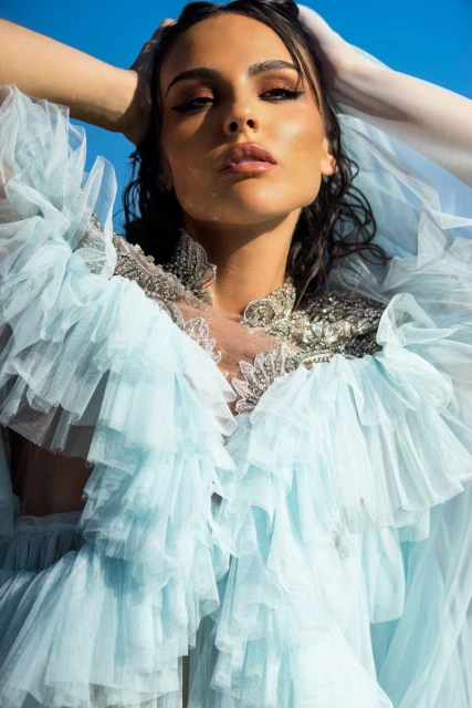 Woman with hands on head, gazing forward, wearing a ruffled light blue gown with decorative beading against a clear blue sky.