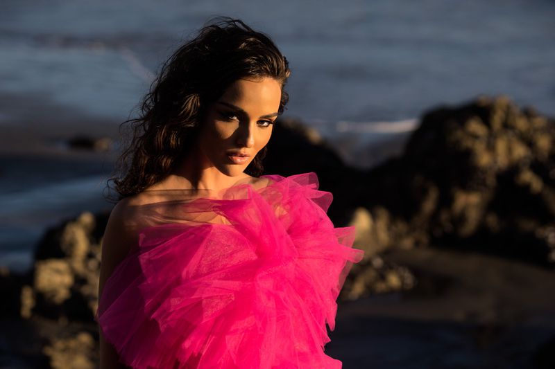Brunette woman wearing a pink tulle dress in an outdoor beach setting with rocks and water creating a natural backdrop.