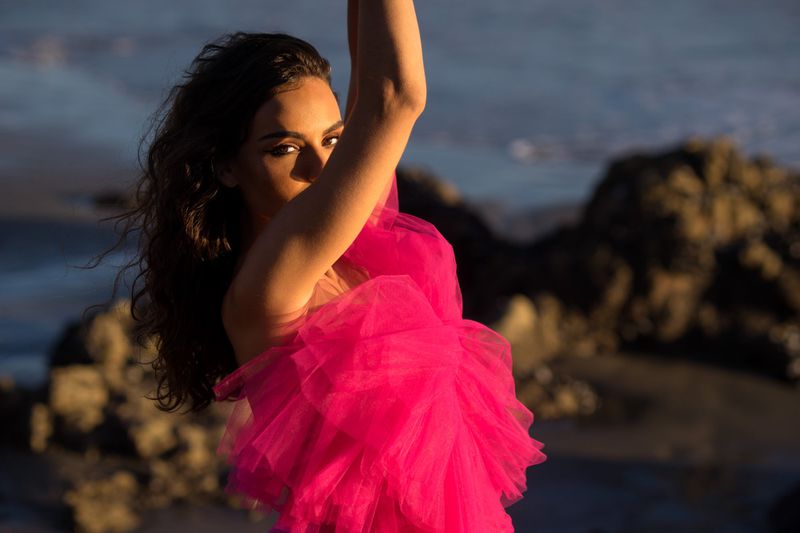 Woman in a vibrant pink tulle dress poses on a beach with dark sand and rocks, creating a striking contrast with the natural setting.