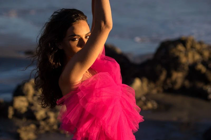 Woman in a vibrant pink tulle dress poses on a beach with dark sand and rocks, creating a striking contrast with the natural setting.