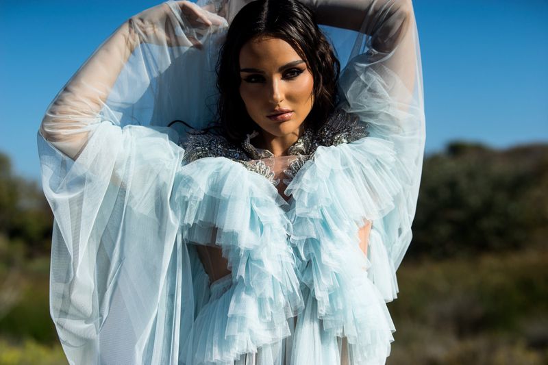 Woman wearing an icy blue ruffled tulle dress with raised arms against a natural landscape and clear sky.