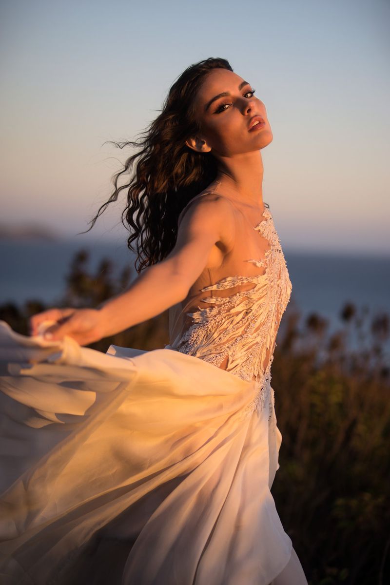 Elegant woman in flowing dress poses outdoors at sunset, showcasing intricate details and soft lighting against a natural backdrop.