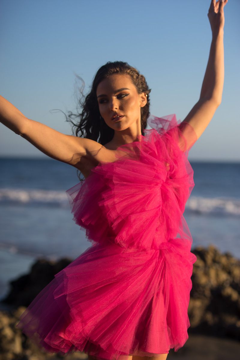 Model posing on a rocky shoreline wearing a vibrant hot pink ruffled tulle mini dress against the ocean.