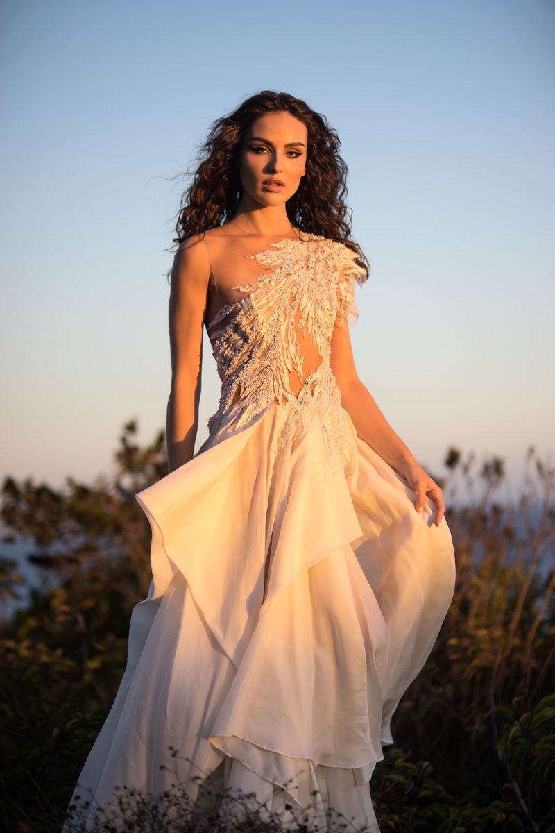 Woman in ornate white gown poses in natural setting against blue sky at dusk. Fashion editorial photo highlighting design and ethereal mood.