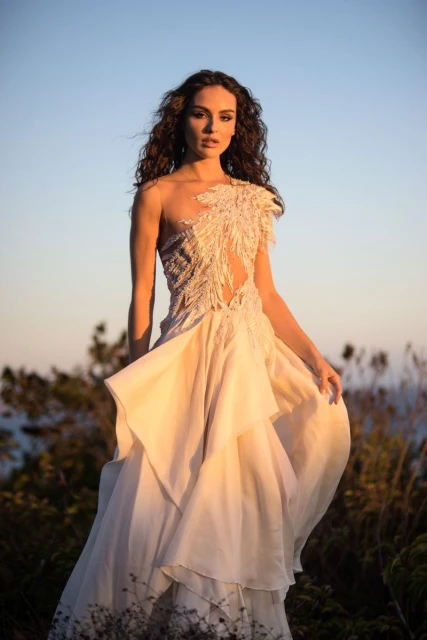Woman in ornate white gown poses in natural setting against blue sky at dusk. Fashion editorial photo highlighting design and ethereal mood.