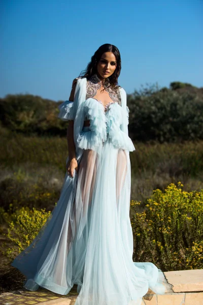 Woman wearing a light blue ruffled evening gown posing outdoors near bushes and blue sky