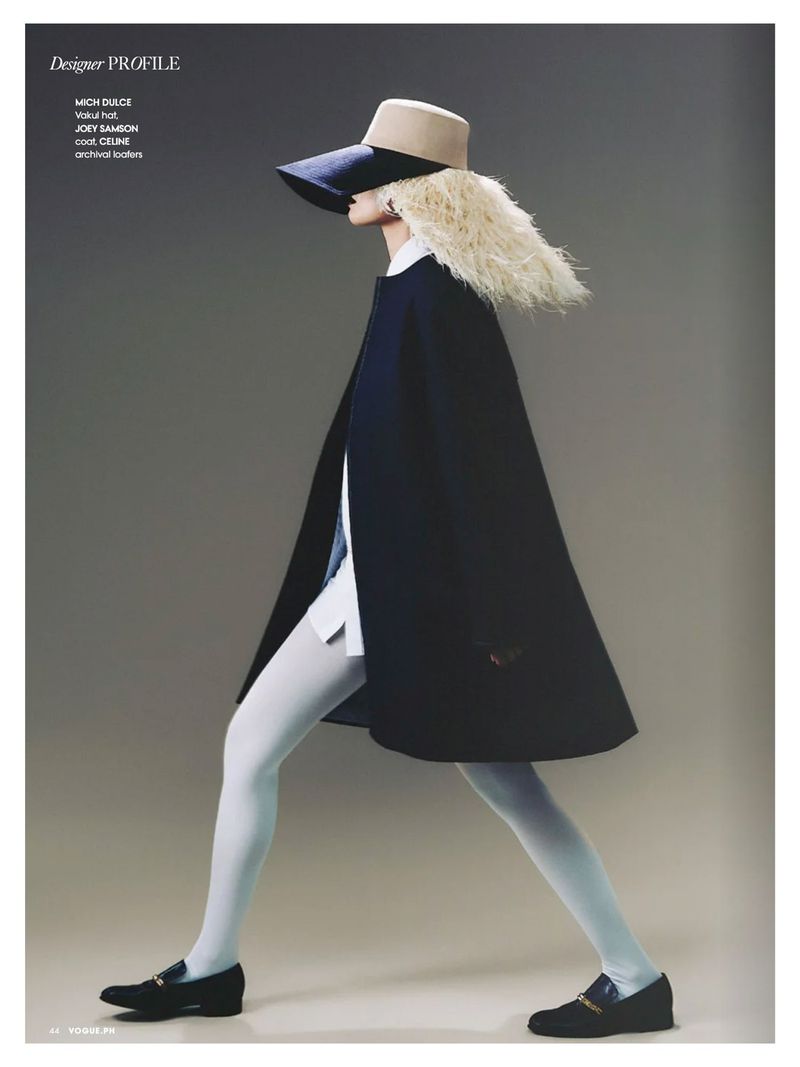 Fashion editorial shot: A woman in a wide-brimmed hat, cape, and tights strides against a gray backdrop, showcasing a stylish, retro-inspired ensemble.
