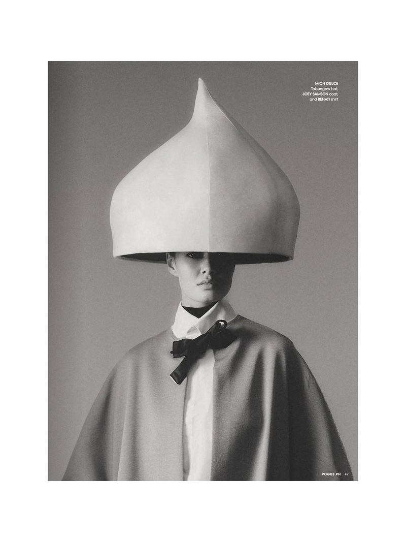 Monochrome fashion portrait of a person wearing a large, unusual cone hat, cape, collared shirt, and bow tie against a gray backdrop.
