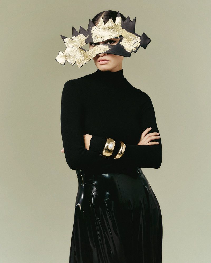 Fashionable woman in black turtleneck and shiny skirt wearing abstract gold and black mask and gold cuff bracelets in studio setting.