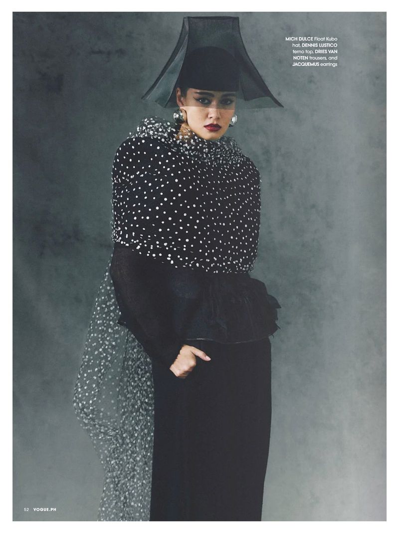 Editorial fashion shot of a woman in a black and white polka dot top and large black hat posing against a dark grey background.