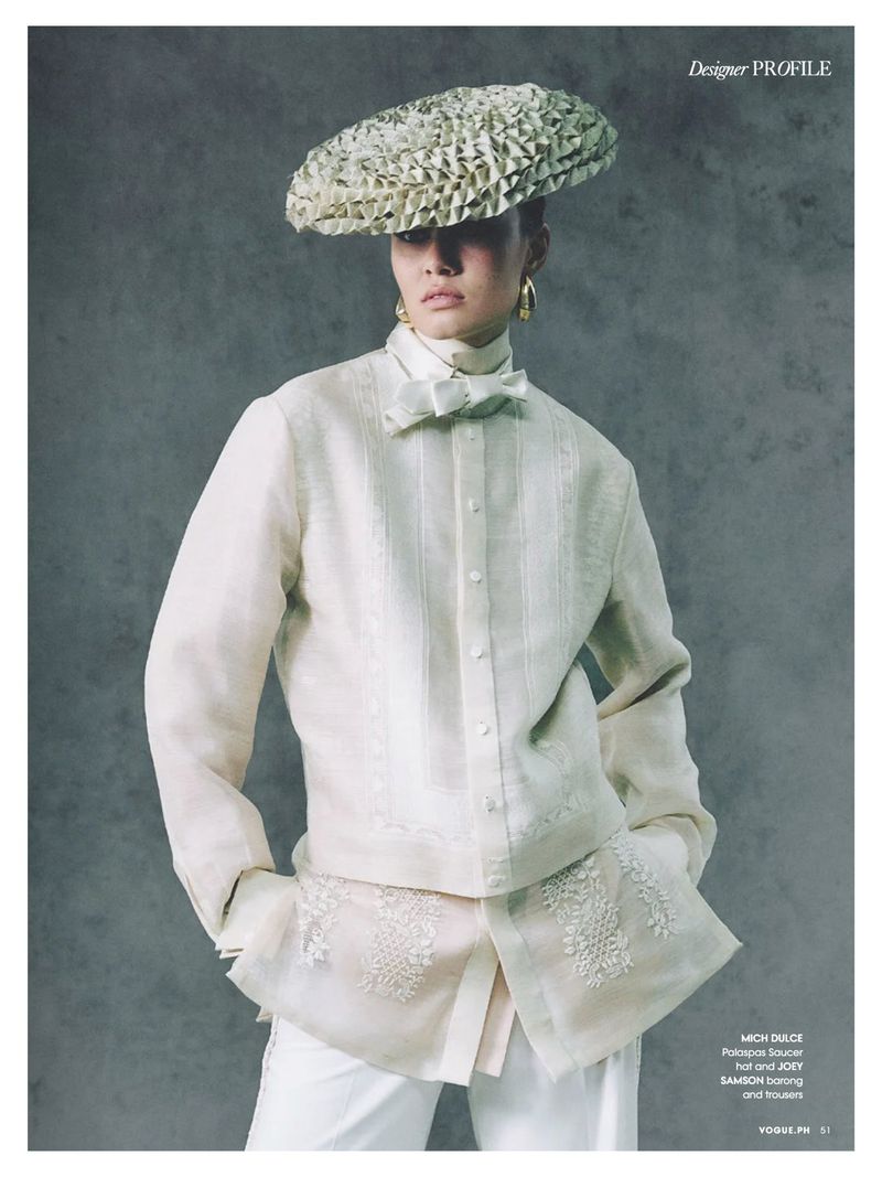 Fashion editorial shot of a person wearing a woven hat, bow tie, and traditional-style shirt with embroidered details on a grey background.