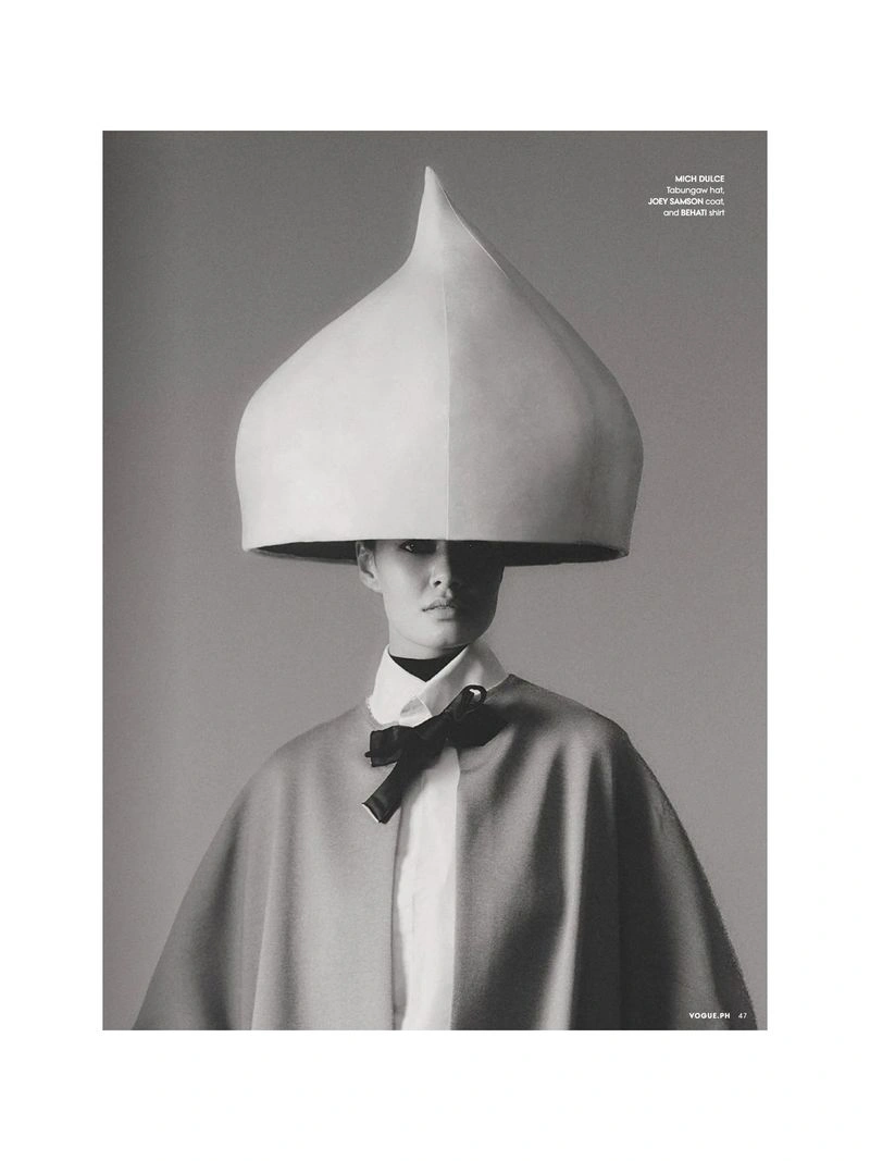 Model wearing an oversized sculptural droplet headpiece, collared shirt with black bow, and draped cape in a black and white portrait.