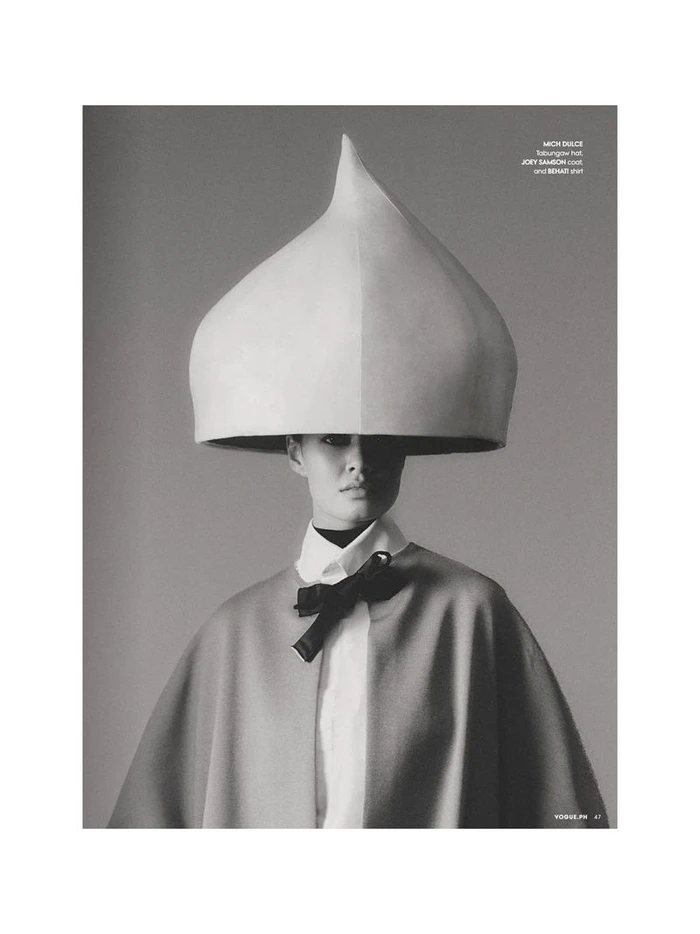 Model wearing an oversized sculptural droplet headpiece, collared shirt with black bow, and draped cape in a black and white portrait.