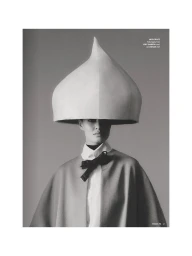 Model wearing an oversized sculptural droplet headpiece, collared shirt with black bow, and draped cape in a black and white portrait.