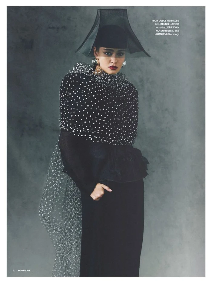Model in black polka dot cape and sculptural sheer hat against a textured grey background for Vogue Philippines editorial.