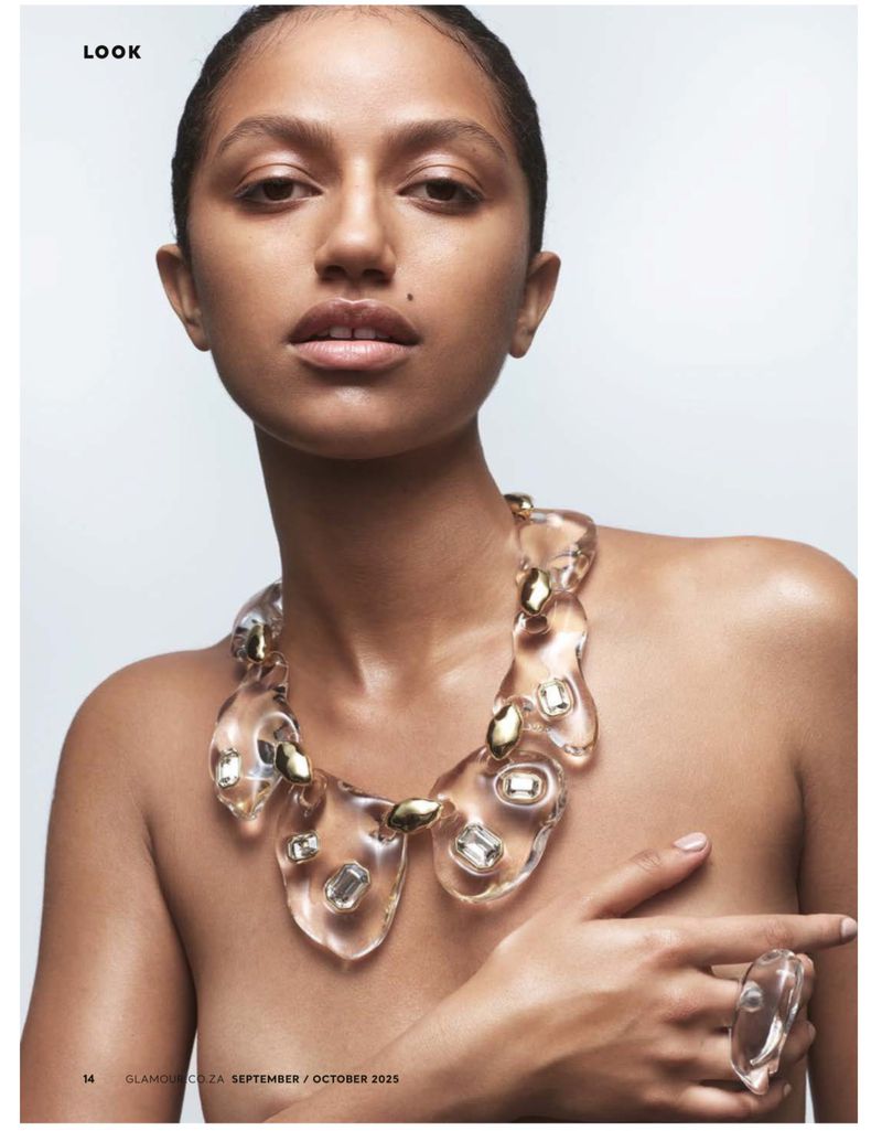 Close-up studio shot of a tan-skinned woman with slicked-back hair wearing a clear statement necklace and ring, posing against a white backdrop.