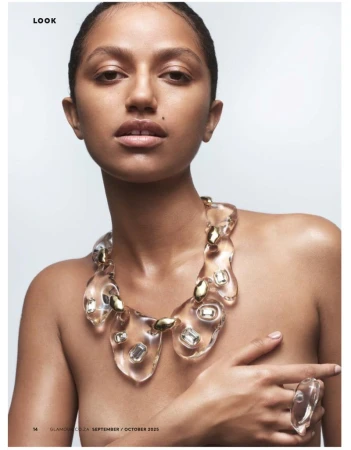 Close-up studio shot of a tan-skinned woman with slicked-back hair wearing a clear statement necklace and ring, posing against a white backdrop.