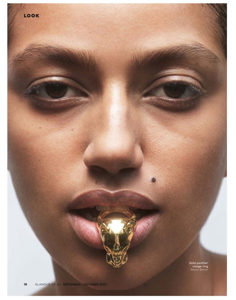 Close-up studio shot of a woman with a gold jaguar ring in her mouth, neutral makeup, against a pale background for a beauty editorial.