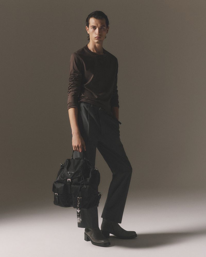 Fashion shot of a slender adult male model with backpack posing in long-sleeve shirt and dark pants in a neutral grey photo studio setting.