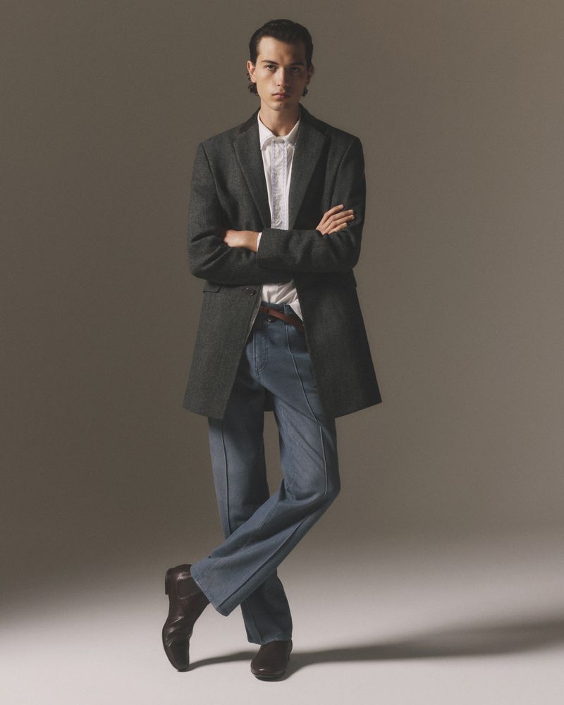 Full-length shot of a young man in a grey wool coat, white shirt, and blue pants, posing indoors with crossed arms and legs.