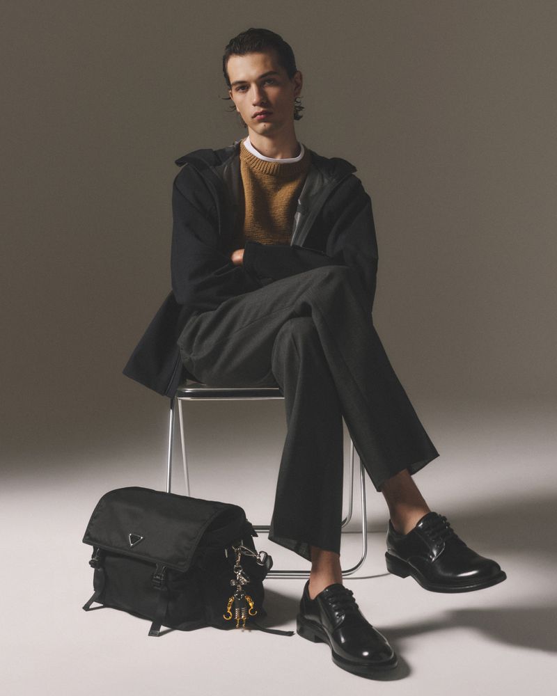 Man in a sweater and jacket seated next to a black bag and wearing black shoes, studio shot with grey background, looking at the camera.
