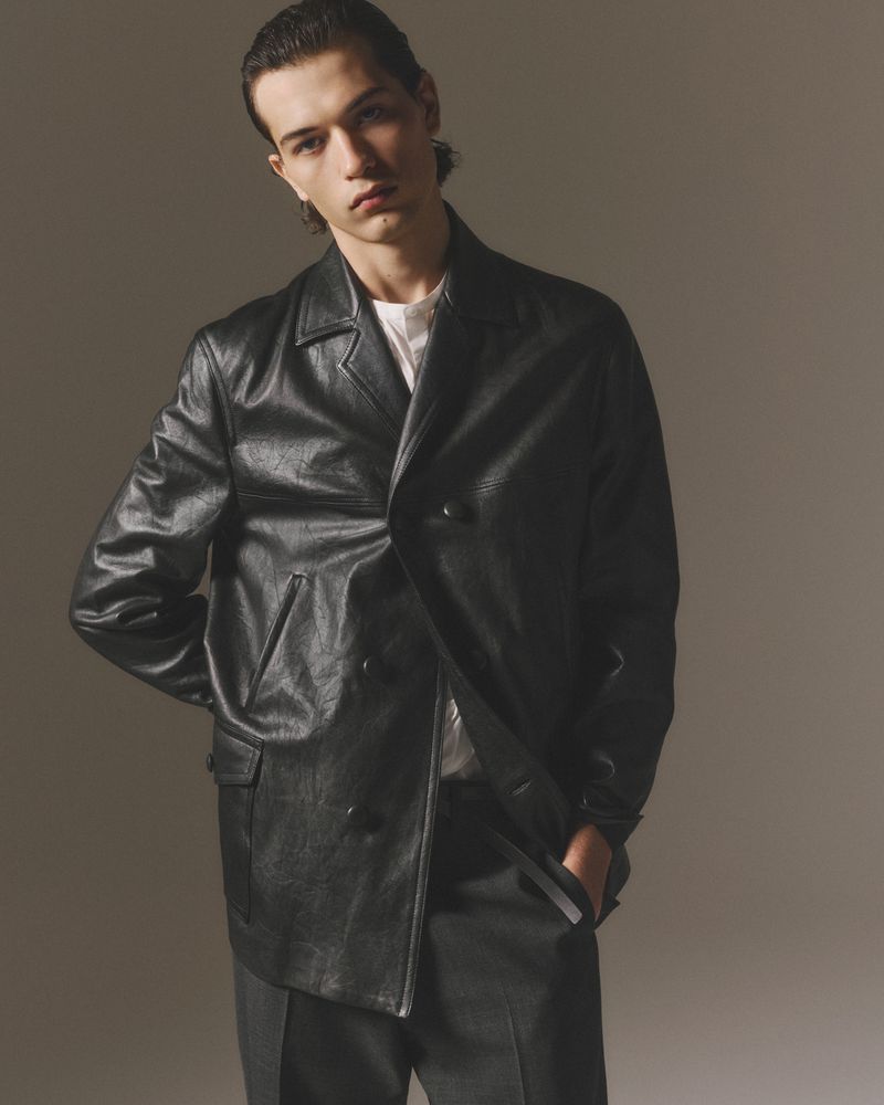 Portrait of a young adult male wearing a black leather jacket and dark pants in a neutral indoor setting with studio lighting.