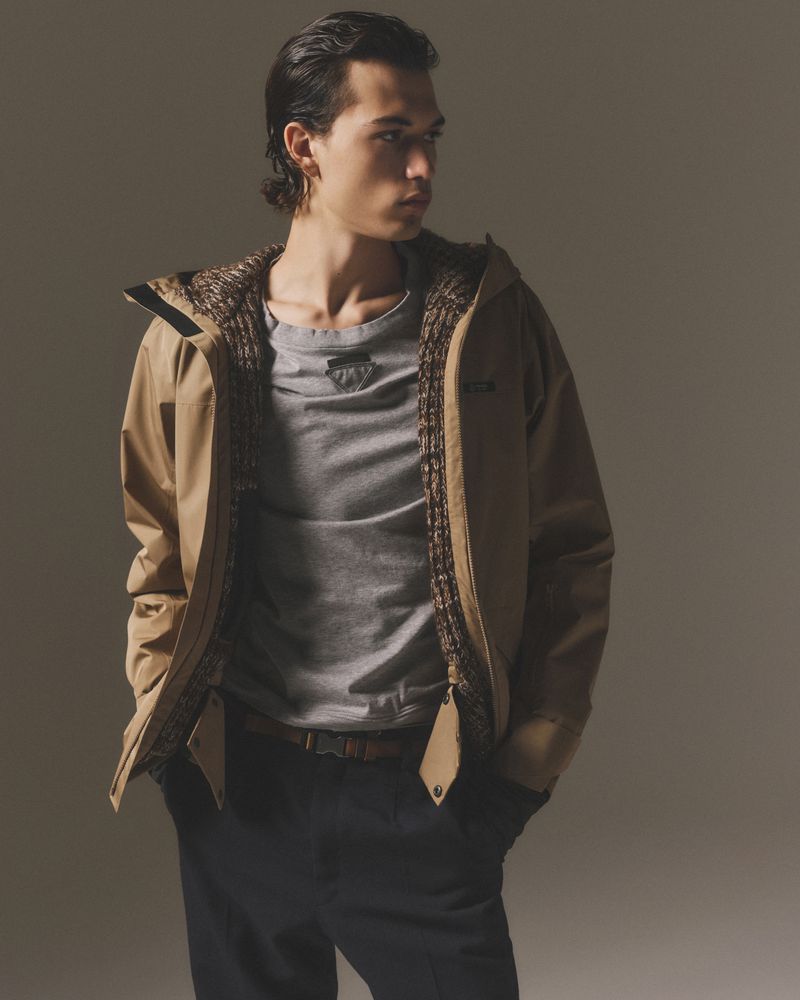 Stylish male model in a tan hooded jacket and grey t-shirt poses indoors in a studio, looking away from the camera with a neutral expression.