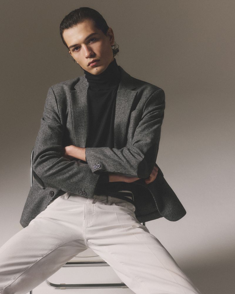 Man in gray blazer and white pants poses indoors. He is sitting on a stool with arms crossed in a fashion-style portrait.