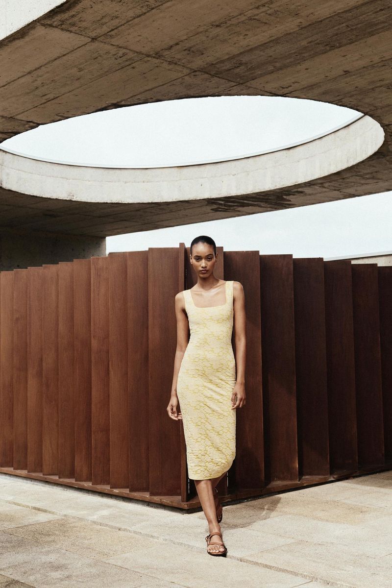 A stylish adult woman stands in a fitted, light-yellow dress and sandals against a modern, geometric, architectural background on a bright day.