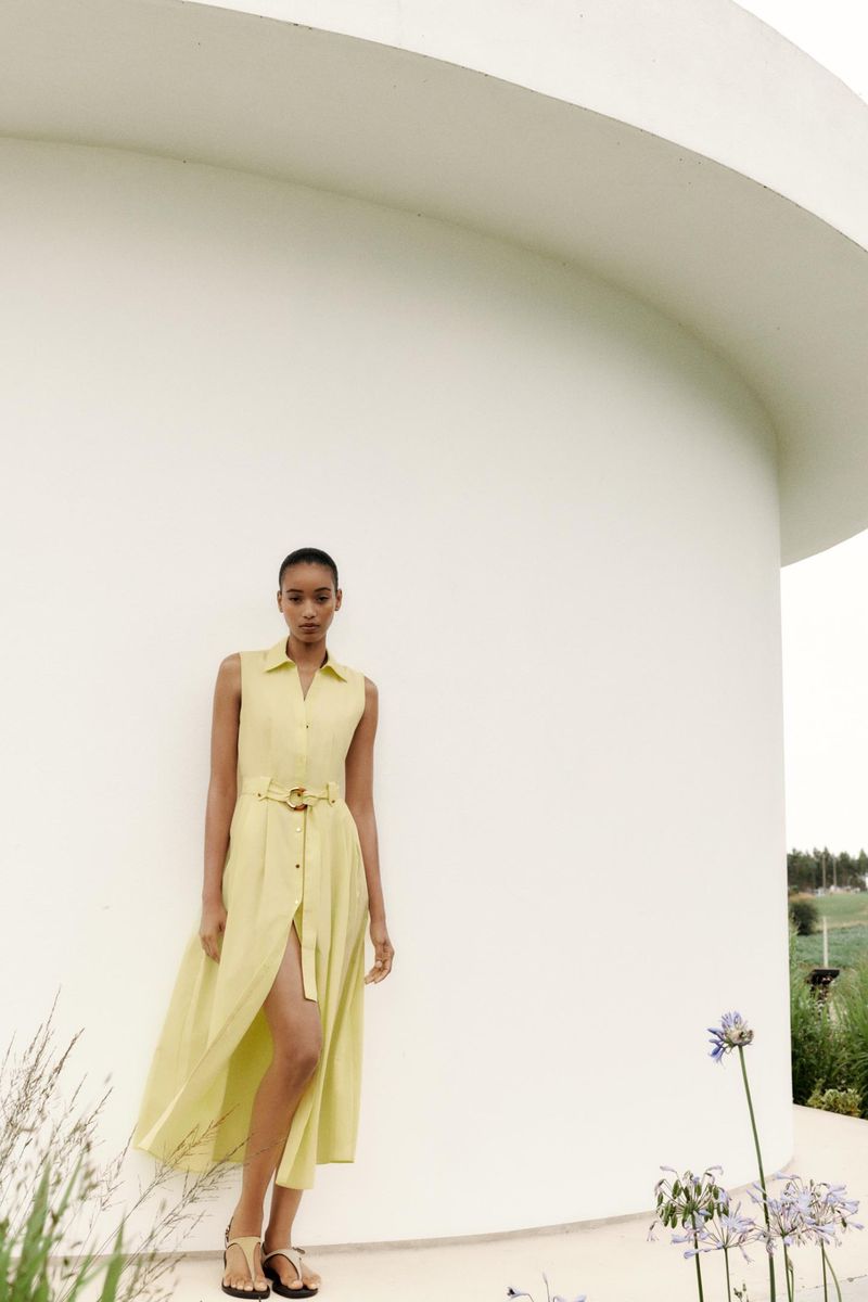 A woman poses against a white building in a yellow dress. She is wearing sandals and has a neutral expression. The background is green.