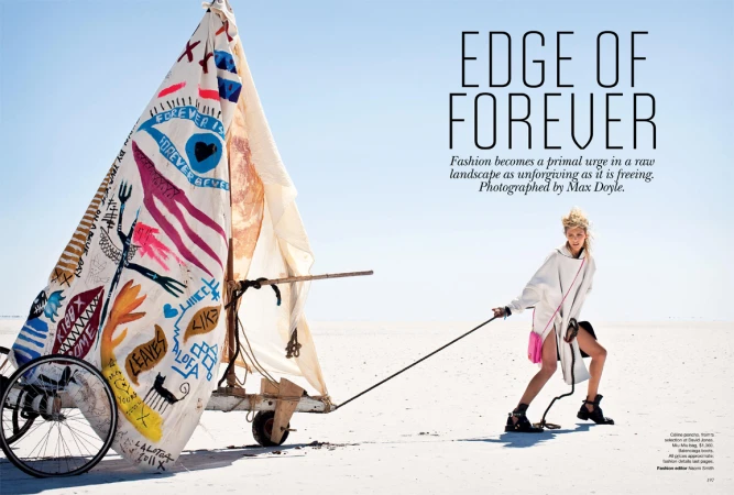 Fashion shot of a woman in a hooded outfit pulling a sail-cart on a salt flat under a bright sky, featuring hand-painted graphics.