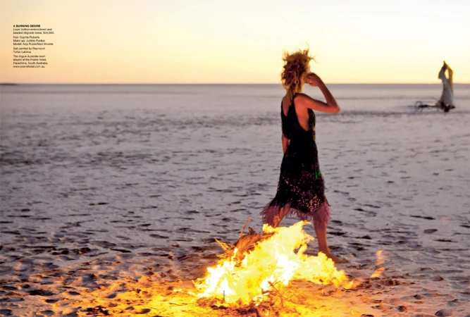 A woman in an evening gown standing in front of a fire on a beach at sunset, with another figure walking away in the distance.