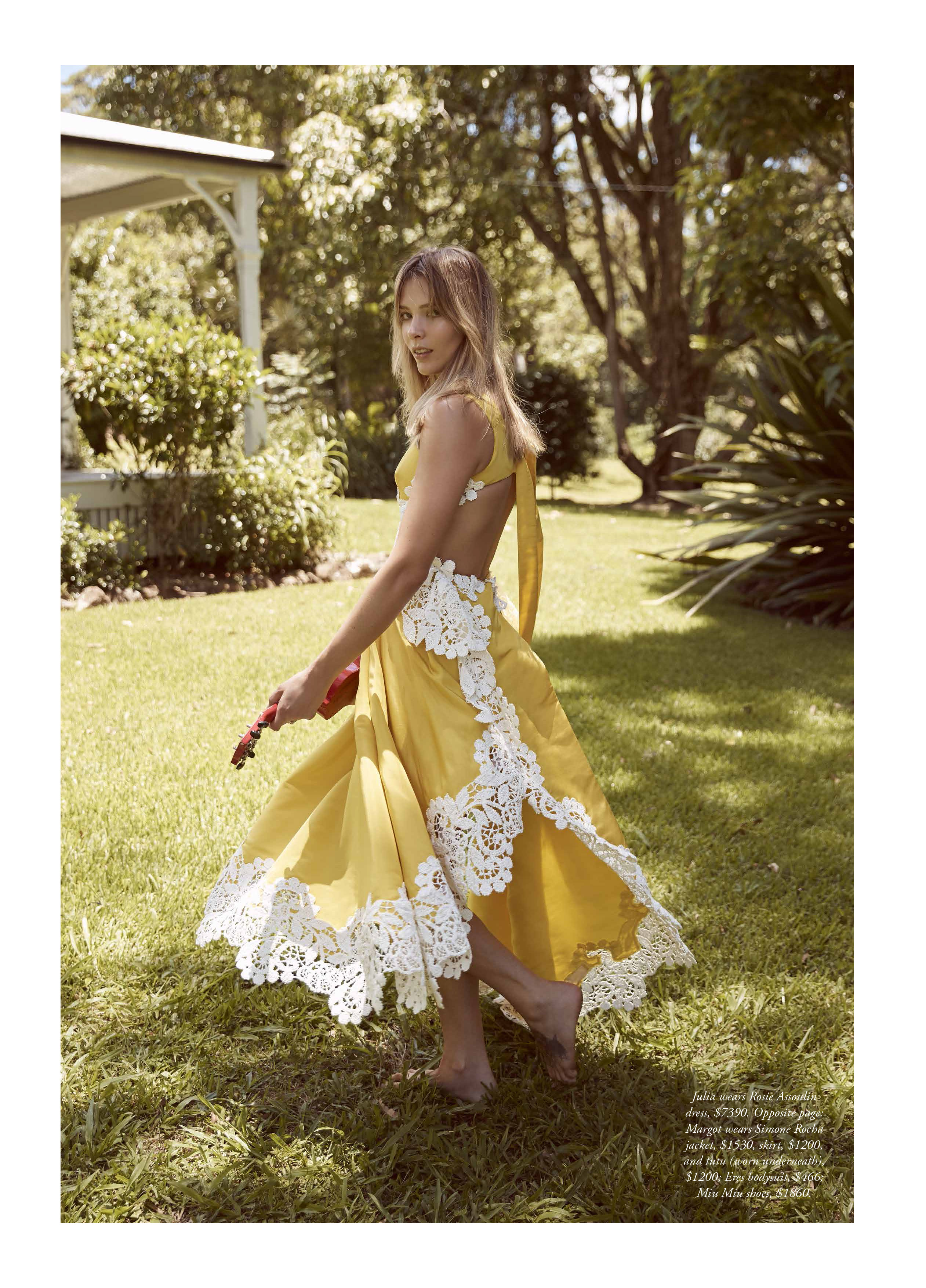 A woman in a yellow dress with lace trim stands in a green lawn, looking over her shoulder toward the camera in soft outdoor light.