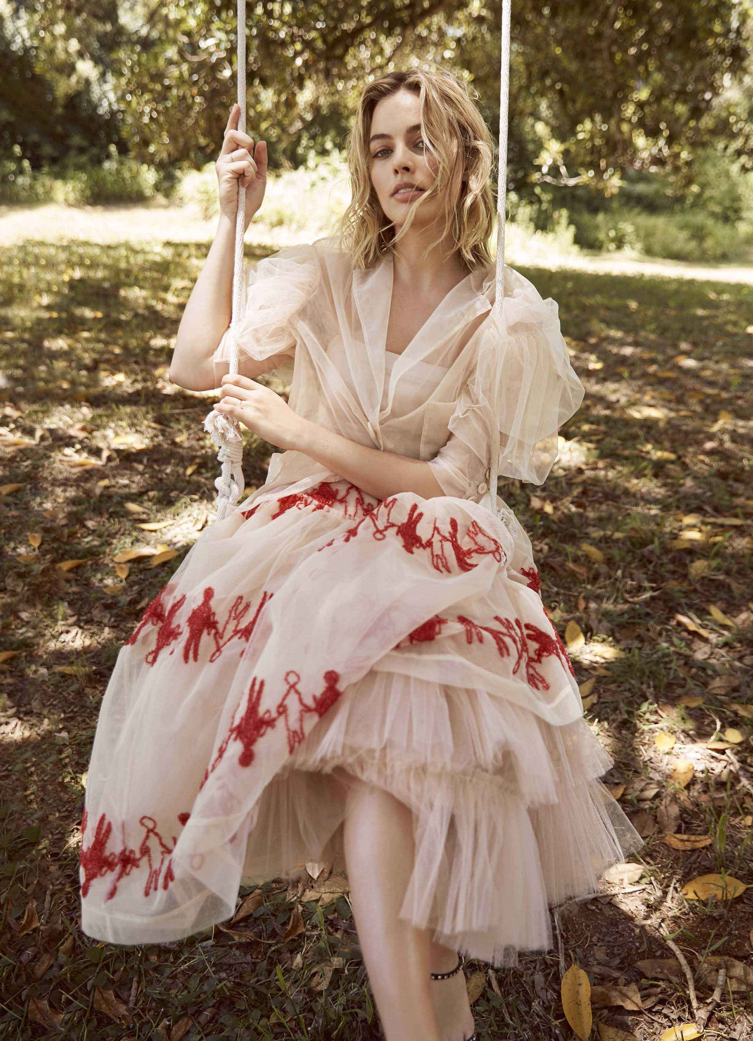 Adult woman in flowing, layered cream-colored dress with red figure pattern seated on rope swing in outdoor leafy setting.