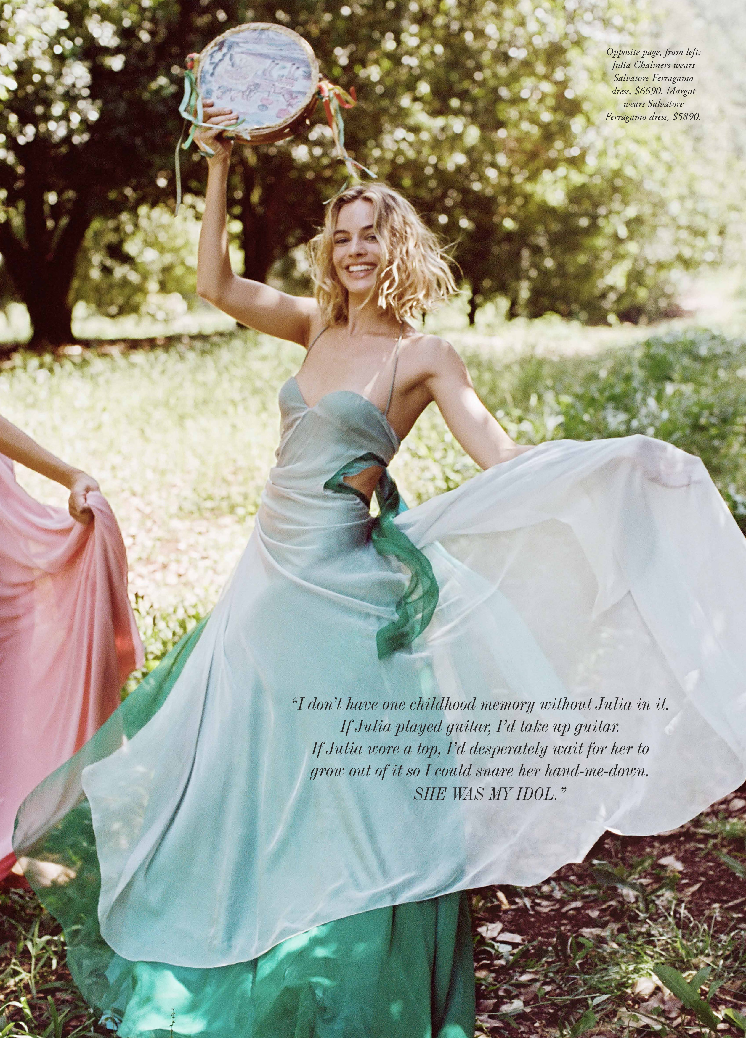 Young woman in flowing dress dances outdoors holding tambourine, smiling, with second woman partially visible in soft, natural lighting.