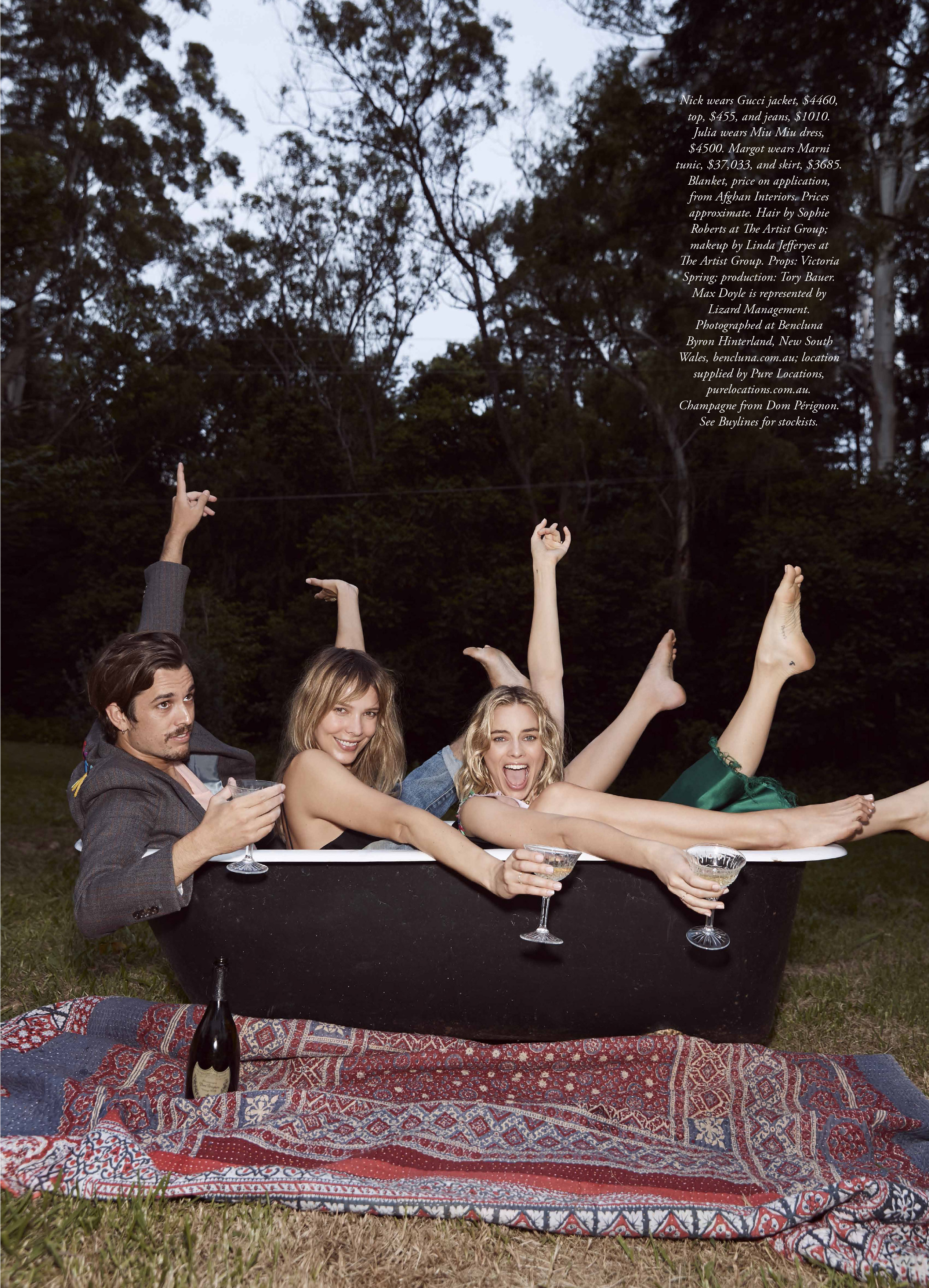 Three friends celebrate in a bathtub outdoors with champagne. Woman in the center smiles with blonde hair, wearing a tank top.