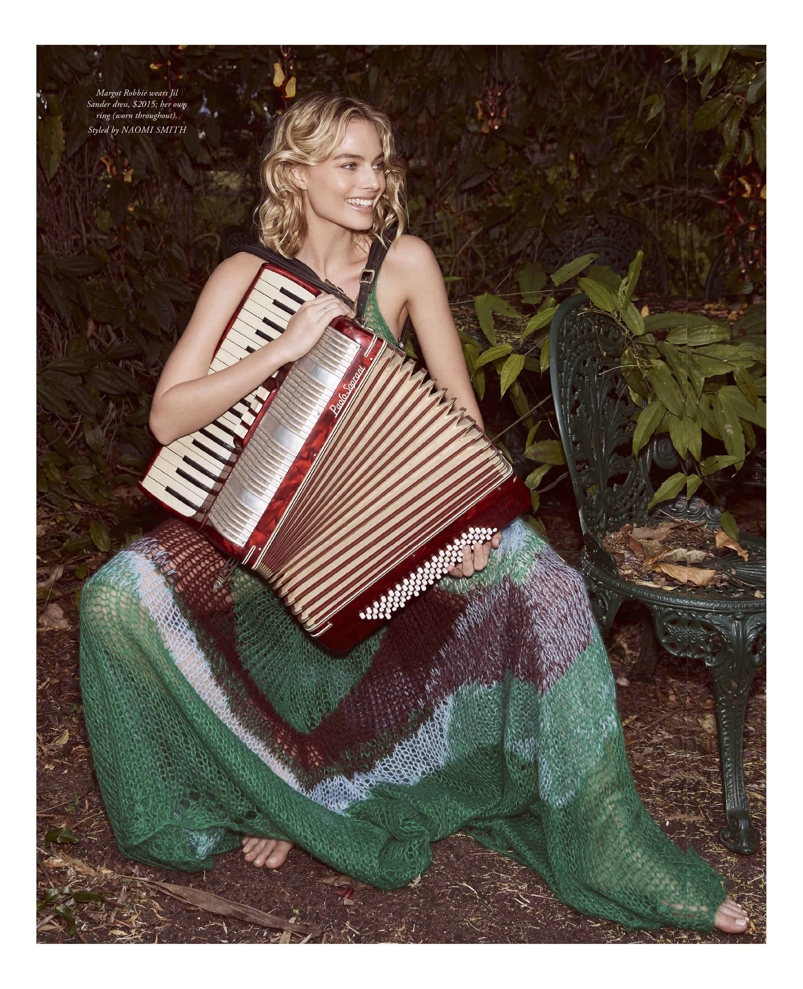 Smiling woman in a long dress sitting outdoors, holding a red accordion, surrounded by greenery in a candid fashion shot.