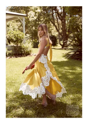 Margot Robbie twirls barefoot on a grassy lawn wearing an open-back yellow dress trimmed with scalloped white lace.